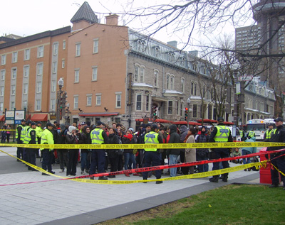 Photo: des policiers portant des dossards jaune encerclent une petite foule. Le tout est entouré de cordons plastifiés jaune et rouge. La moitée de la rue est bloquée. On voit derrière, environ 500 mètres plus loin, le Complexe G.