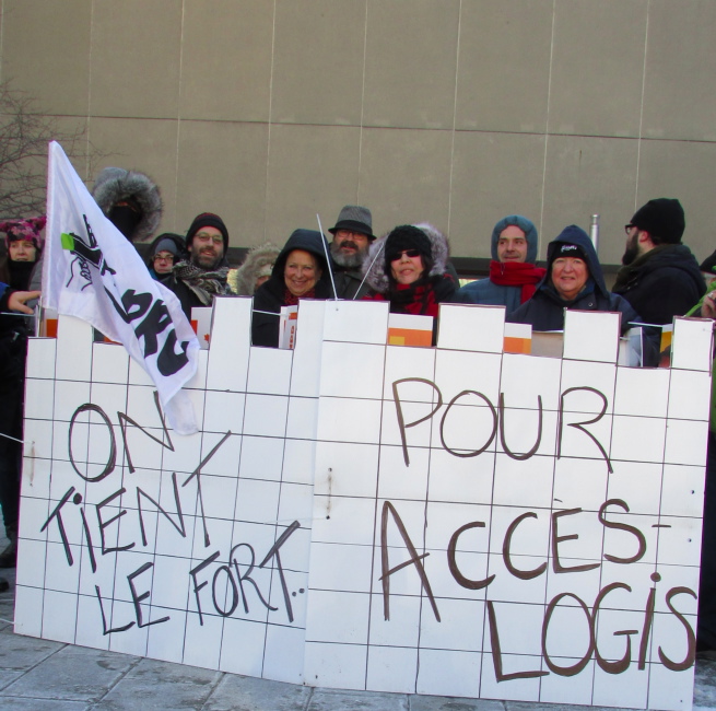 Photo : plus d'une dizaine de personnes derrière un faux fort en carton blanc avec les mots : On tient le fort  ... pour AccèsLogis. Drapeau du FRAPRU.