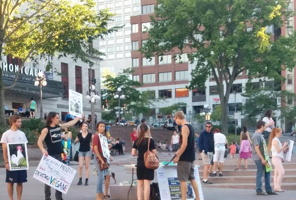 Photo : journée ensoleillée sur la Place d'Youville.  Une dizaine de jeunes personnes discutent et portent des pancartes. D'autres s'amusent sur la place comme à la normale. Arbres verts derrière et Palais Montcalm.