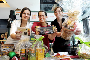 Photo : dans une cuisine, trois jeunes femmes (toutes portant des lunettes) tiennent des aliments emballés. La table devant est bourrée de choses comestibles, souvent emballées pour Noël.