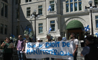 Photo : devant l'Hôtel de Ville, des employés et membres de groupes en défense de droits tiennent une bannière blanche : Pour le droit de manifester à Québec. Marie-Ève Duchesne parle au micro.