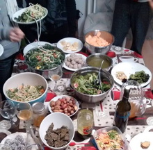 Photo : hormis quelques jambes, on voit une table couverte de nombreux plats : diverses salades, noix, lasagne végé, vin, etc.