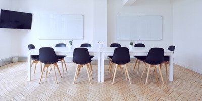 Photo : grande salle blanche propre avec deux grands tableaux blancs d'école. Il y a dix petites chaises de couleur noir autour de tables blanches ainsi qu'un écran télévisé géant suspendu dans le coin.