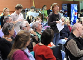 Photo d'une salle comble en 2011 lors d'une consultation qui allait transformer les Conseils de quartier et Conseils de districts électoraux. Une dame parle au micro des citoyens/citoyennes.