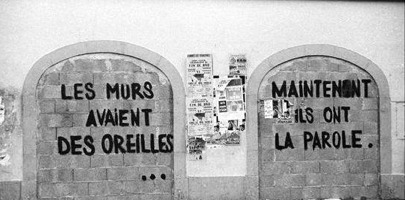 Photo en noir et blanc : graffiti en grosses lettres noires se lisant « Les murs avaient des oreilles. Maintenant ils ont la parole ». Sur un ancien mur extérieur de plâtre, sur la brique qui bloque deux anciennes entrées de rue. 