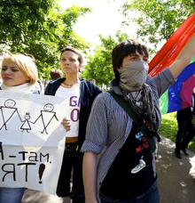Photo lors de la fierté gaie à Moscou (Moscow Pride). On voit trois femmes tenant affiche et drapeau. Celle la plus en avant est jeune, aux cheveux courts bruns, et porte un foulard pour se masquer le visage.