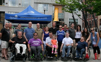 Photo de groupe sur le côté du parvis de l'église Saint-Roch : cinq hommes en fauteuil roulant, treize personnes derrière. Chapiteau bleu de la Ville de Québec.