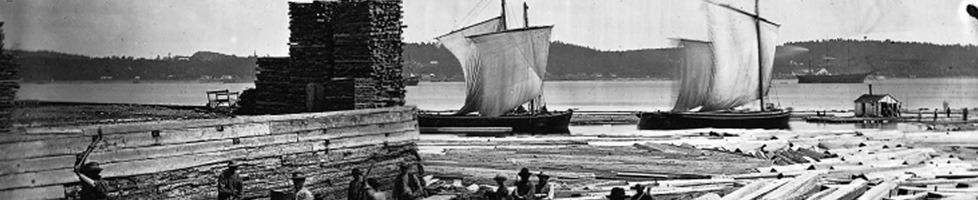 Photo ancienne (noir/blanc) : sur une large rivière ou fleuve, deux bâteaux à voiles blanches tout près d'un port où l'eau contient du bois flottant.  Un bâtiment de bois est aussi en construction.