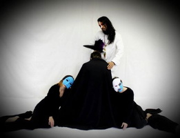 Photo conceptuel pour la pièce : trois personnes portant des capes noires et des masques (blanc et bleu), écrasées au sol une contre l'autre, l'air morose, devant un homme en blanc aux cheveux longs bruns.