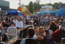 Photo d'une centaine de gens sur des chaises extérieures. Jeux gonflables bleu ciel, glissade arc-en-ciel et tente bleue marine de la Ville.