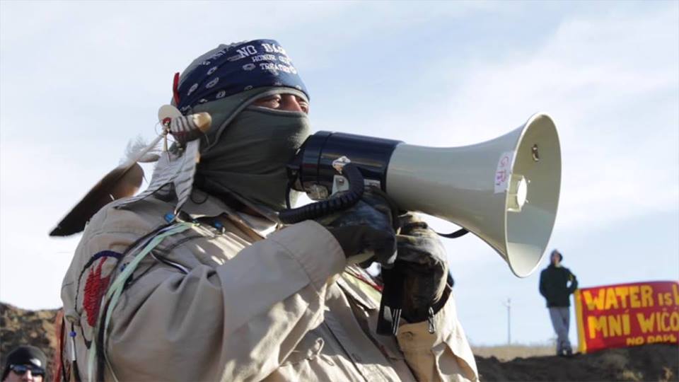 Photo : un homme autochtone, probablement âgé, parle dans un porte-voix.  Son visage est masqué et il porte un foulard sur la tête avec les mots « No Black Snake - Honor... », ainsi que des plumes et symboles de sa nation.