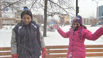 Photo d'un reportage de TVA : garçon et fillette, peau brune, devant un parc et il neige. Manteau noir et gris et long manteau rose pour la fille. L'air content.