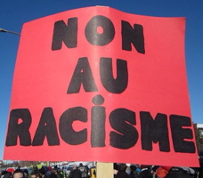 Photo d'une pancarte rouge lors d'une manif : NON AU RACISME. On discerne une foule derrière.