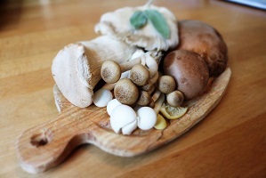 Photo: plusieurs types de champignons comestibles placés serrés sur un mini-plateau en bois en forme de noisette, sur une table en bois.