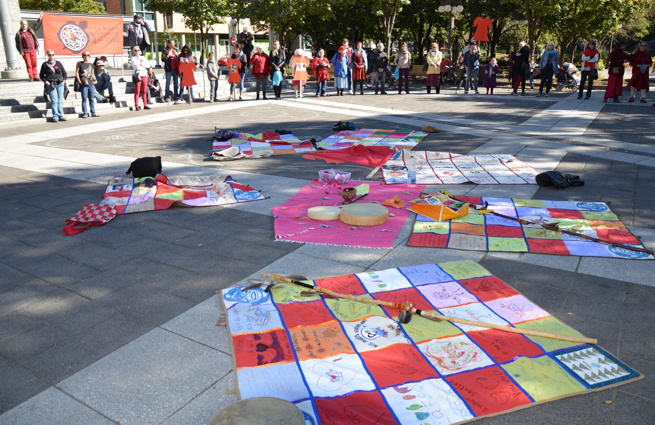 Photo : cinq couvertures courte-pointe (lire: mosaïques de carrés colorés) sur lesquelles sont écrits des messages; tambourins au milieu ; on voit une partie de la foule autour, dans un grand cercle sur la Place de L'Université-du-Québec.