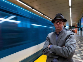 Photo : Jason Prince à côté d'un train de métro bleu qui passe rapidement. Chapeau style 1950 gris foncé, lunettes à bordures noires, veston bleu-gris, bras croisés.