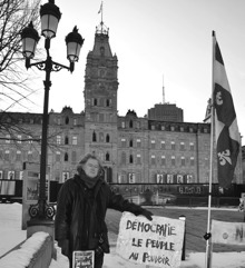 Photo par Réal Michaud en noir et blanc : grand manteau de cuir noir, cheveux longs, la main sur une affiche « Démocratie - Le peuple au pouvoir », drapeau du Québec et, derrière, la façade de l'Assemblée nationale du Québec.