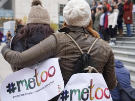 Photo : deux jeunes femmes, de dos, une soutient l'autre moralement avec son bras son ses épaules. Elles portent sur leur dos des affichettes colorées « #metoo ». Plus loin, des femmes se tiennent unies sur des marches publiques de ciment.  C'est probablement à Vancouver considérant la source sur Vice News.