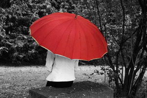 Photo en noir et blanc : une dame est assise, de dos, sur un siège carré de pierre, dans un parc entouré de verdures. Elle est cachée par un parapluie rouge (seule couleur sur la photo).