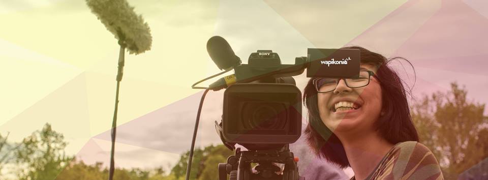 Photo panoramique : une jeune femme très souriante manie une caméra de cinéma. Paysage naturel flou derrière.