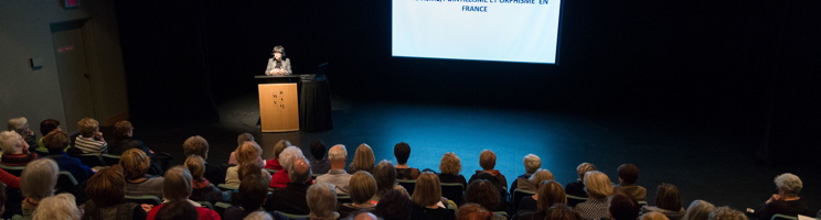 Photo panoramique d'un amphithéâtre où Pascale Mathé donne une conférence avec un écran géant. La salle est comble de gens.