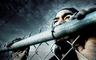 Photo : un homme noir, vu de près, à travers une clôture de métal. L'angle présente aussi le ciel nuageux.