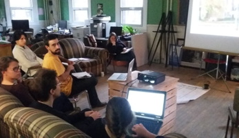 Photo : on voit six personnes dans une grande salle sur des divans, devant un écran blanc sur lequel un schéma est projeté.