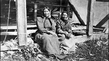 Photo noir/blanc ancienne de deux femmes autochtones assises sur le bord d'une structure en bois carré. Elles portent des robes occidentales d'époque.