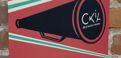 Photo d'une affiche sur un mur de briques rouges intérieures. Un cône noir, soit un porte-voix, et le bout affiche « CKIA 88,3 radio urbaine ». Le fond est rouge, avec deux lignes de couleur vert pâle, en diagonal, en-dessous et au-dessus.