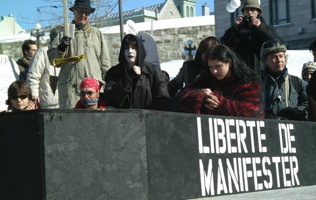 Photo : devant un cercueil noir, sur lequel est écrit « Liberté de manifester ». Une personne, au visage peinturé en blanc, joue la mort. Plusieurs autres autour sont les personnes endeuillées. Un jeune homme a la bouche baillonnée. Un monsieur tient une chandelle.