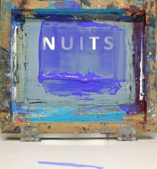 Photo d'une des oeuvres qui ressemble à un écran de télévision, mais peinturé de plusieurs couleurs ici et là, sauf le mot NUITS est écrit clairement en lettres blanches et droites. Sur la table devant, une trait de peinture bleu-mauve.
