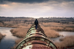 Photo : un homme, de dos, marche sur un gros pipeline d'oré qui passe sur un grand champ marécageux. L'herbe est beige. L'homme n'est pas un travailleur selon son style vestimentaire normal.