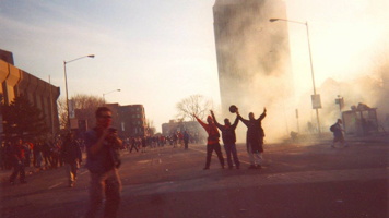 Photo : l'édifice Complexe G est couvert de gaz lacrymogène.  Trois jeunes hommes, les bras levés comme en victoire, devant. Un photographe est aussi dans la photo. Au loin, une foule de gens sur la rue.