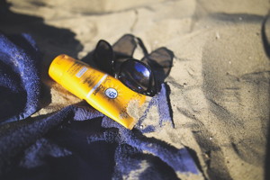 Photo d'un tube jaune sur une serviette bleu avec des lunettes de soleil, sur du sable de plage.