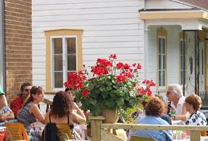 Une dizaine de personnes sur la terrasse au soleil, de style café, sur le devant d'une ancienne église et à côté d'une maison blanche.