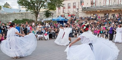 Trois danseuses en grande robe blanche, tenue en quelque sorte en éventail. Une agit plus comme cheffe d'orchestre. Foule  dense et colorée de gens sur les marches devant le Palais Montcalm.