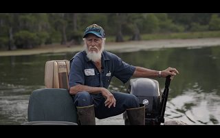 Capture-écran d'une des scène : un homme âgé, l'air sérieux et vénérable, à la barbe blanche et casquette et vêtements bleutée, pilote un petit bateau à moteur sur un lac sauvage.