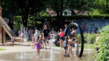 Photo de 2017 : jeux d'eau, enfants dedans, parents autour, glissade, verdure.