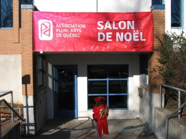 Photo de l'entrée : grande bannière rose: Salon de Noël / Asso. Pluri-Arts de Québec. Mur blanc; cadres de porte et fenêtre bleu.