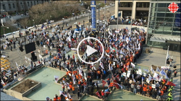 [PHOTO: capture-écran d'un vidéo de Radio-Canada. Vue du dessus de la foule au bord du Centre des congrès de Québec. Foule assez dense d'environ 400 personnes. Beaucoup de petits drapeaux syndicaux.]