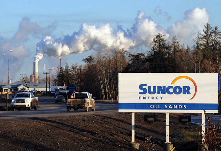 [Desc.: photo d'une affiche routière Suncor au bord d'une route et derrière des cheminées avec des colonnes de fumée.]