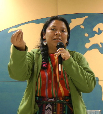 photo: Lolita Chavez, debout, micro à la main gauche, les doigts de sa main droite tenues ensemble pendant qu'elle s'exprime sur comment son peuple voit la vie. Derrière elle, une carte bleu du monde de la salle de conférence de l'Auberge L'Autre Jardin.