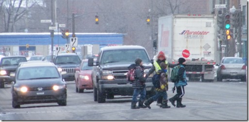 Photo : en hiver sur une grande intersection, des enfants traversent à l'aide d'une employée à la circulation. Plusieurs voitures attendent.
