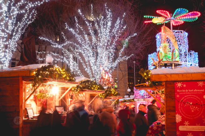 Photo de soir : arbres remplis de lumières blanches ; cabanes en bois bien éclairées ; affiche rouge du Marché de Noël allemand