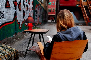 Photo : une jeune femme de dos, sur une chaise, lisant un livre dans une ruelle aux murs pleins de beaux graffitis colorés vert et rouge. Ses cheveux sont roux longs, chandail à capuchon et manteau de cuir noir par-dessus. Petite table de lecture avec une lampe rouge dans la ruelle.