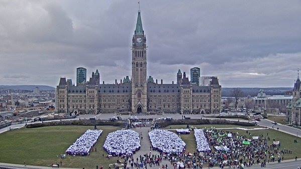 [Desc.: Photo de haut du parlement canadien. Une foule dense forme : 100 %. Ciel bleuté nuageux.]