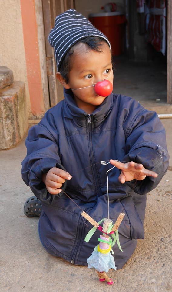 [ Belle photo d'un enfant, portant un nez rouge de clown, et jouant avec une marionnette fabriquée à la main. Le sol est de terre glaise. L’enfant porte une tuque rayée noire/blanche et un grand manteau d’hiver bleu-mauve (donc probablement dans une région montagneuse de l’Amérique du Sud).]