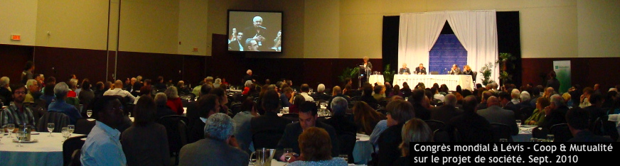 Congres des coop et mutualité sur le projet de société, sept. 2010 - Photo où on voit la grande salle de conférence remplie de gens assis à des tables, avec un écran géant montrant un homme parlant au micro [Palais des congrès de Lévis]