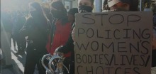 Photo : des jeunes femmes masquées manifestent. Une tient une pancarte se lisant Stop policing women's bodies / lives / choices.