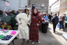 Photo : au soleil derrière l'église, deux participantes posent, avec des expressions exagérées et comiques, en robe de chambre. Plusieurs personnes derrière s'affairent aux tables.